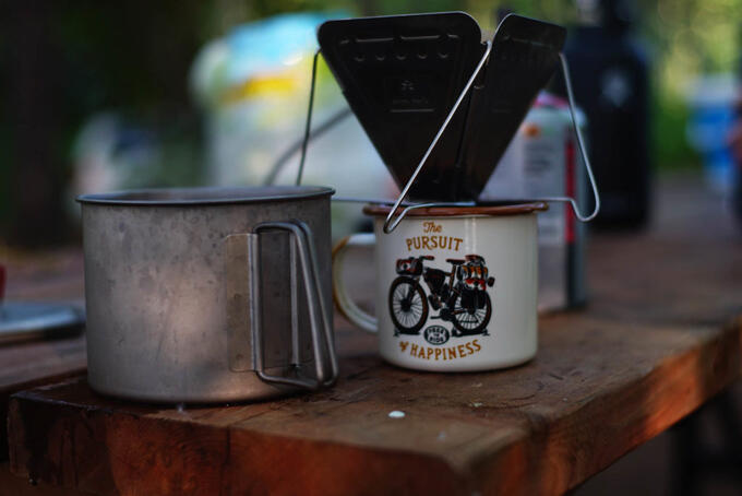 camp supplies and tin cups on a wooden picnic table; one cup sports an image of a bike and reads "the pursuit of happiness"
