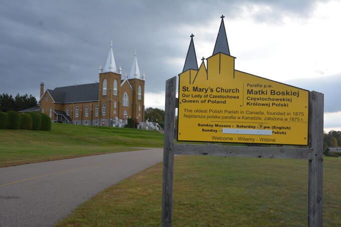 old church in background against blue-grey clouds, yellow church-shaped sign in front reads "St. Mary's Church-- Queen of Poland" in English, Polish and Kashubian