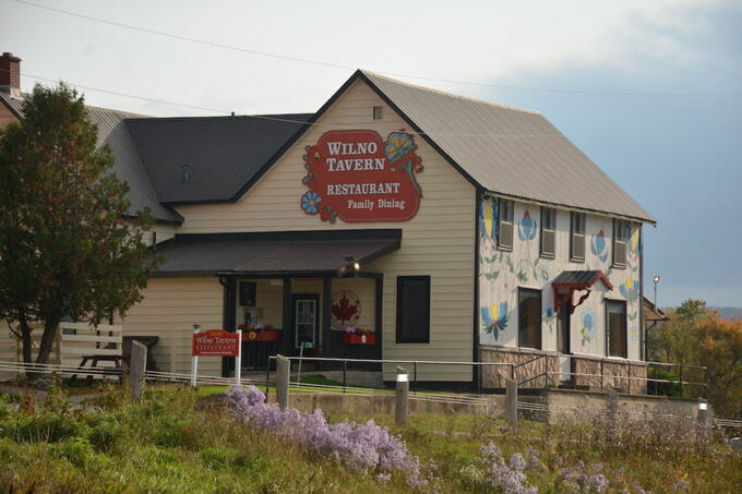 beige building surrounded in trees and flowers and painted with large floral motifs, sign on front reads "Wilno Tavern Restaurant and Family Dining"