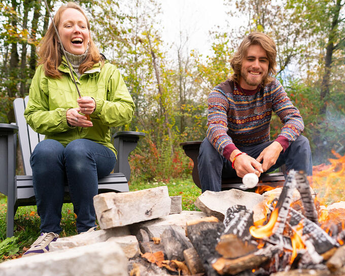 people in jackets and sweaters laughing around campfire with forest in background