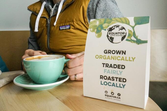 person holding a large teal cup and saucer, sitting at a wooden table with a bag of organic fair trade coffee on their left