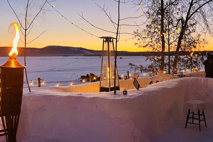 outdoor bar made out of snow, decorated with tiki torches and lights, with frozen lake and orange sunset in background