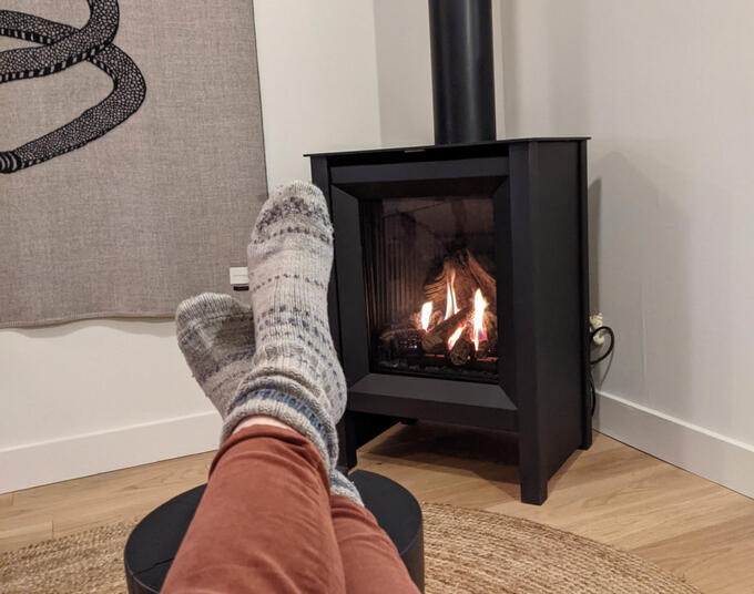 person's feet wearing knitted socks, resting on a stool in front of blazing wood stove
