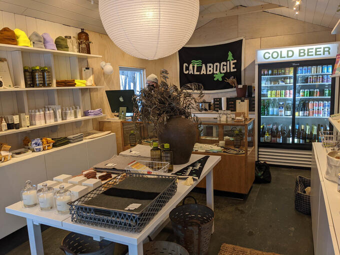 small shop interior featuring rows of items such as soaps, lotions, gifts, toques, a drink cooler, and a flag that reads "Calabogie"