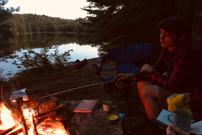 person in red plaid jacket roasting marshmallow over fire next to lake at twilight