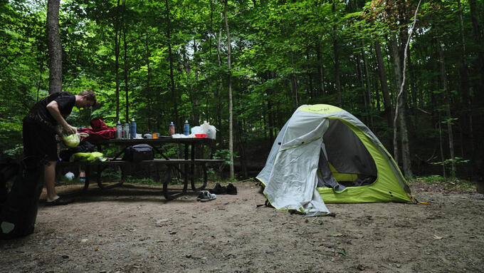 person tending a campsite with green tent and wooden picnic table surrounded by lush green forest