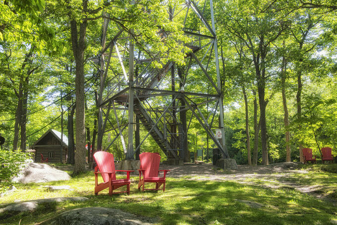 Two red Adirondack chairs are shown in a forested setting.