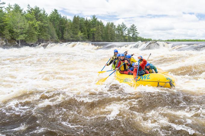 A group of people wearing lifejackets and helmets in a yellow raft in the middle of whitewater