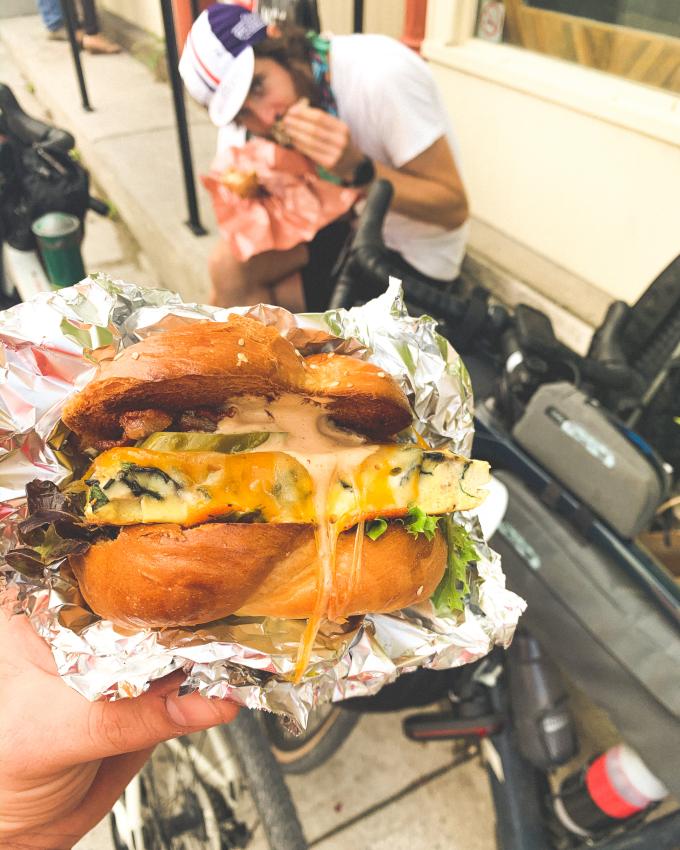 A hand holds a burger in the foreground, and another man in the background eats seated on a bench