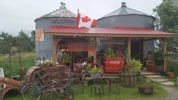 a tin-roofed booth in front of two metal granaries with a wooden sign that reads "General Store"; the booth is full of plants, chairs, antique bicycles and farm equipment, and other paraphernalia. A field and forest are in the background.