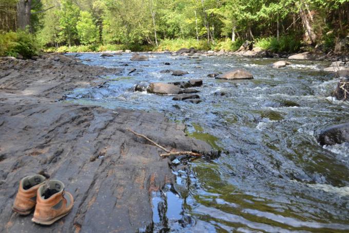 a rocky river with slick rock banks, surrounded by green forest; a pair of brown leather shoes sit on the bank to the left