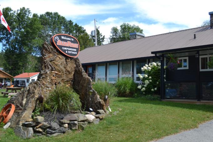 a brown building surrounded by plants, flowers and green grass, and a large tree stump adorned with a sign that reads "Welcome to Bear Ridge Campground and Cottages"