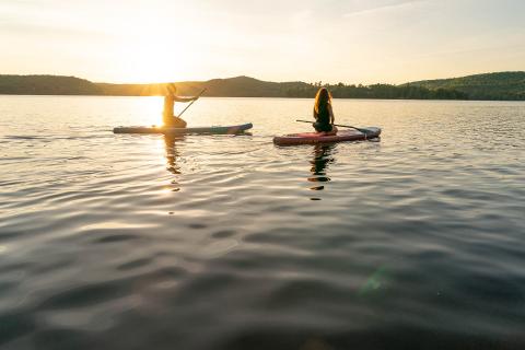 two people kneeling on paddle boards floating on lake with sunset in background