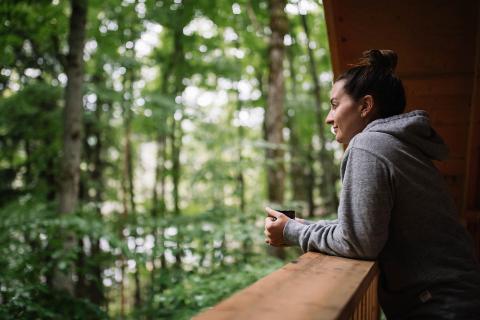 Person smiling and leaning on wooden deck railing, looking at lush green forest