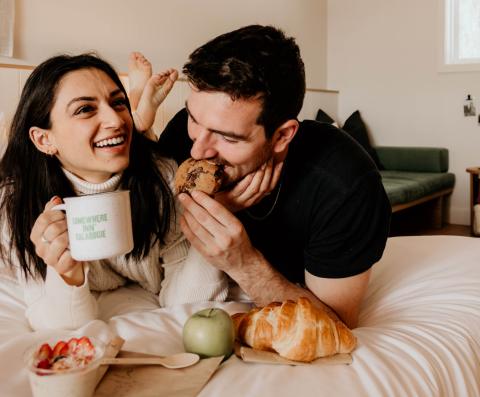 couple laughing and cuddling on bed while enjoying baked goods, coffee and fruit