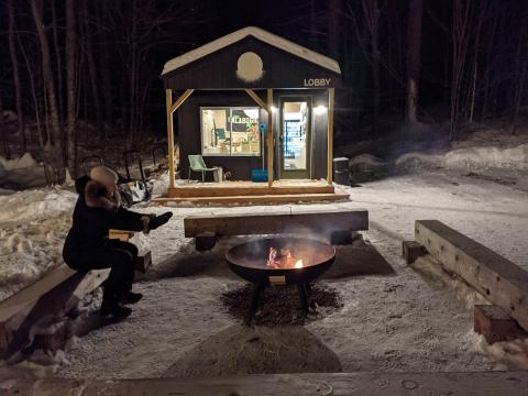 smiling person in winter gear warming hands by outdoor fire at night; small wooden building in background has lit windows and a sign that reads "Lobby"