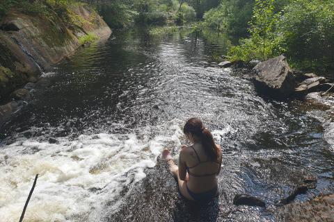 person in bathing suit sitting on a rock in a river surrounded by lush green trees on a sunny day