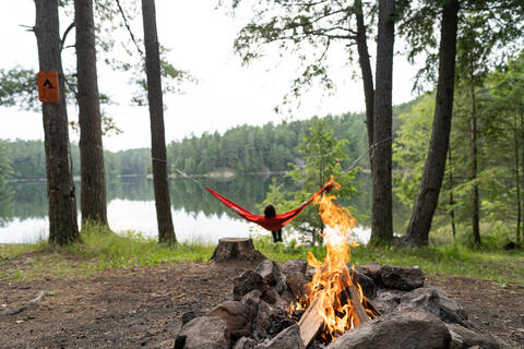 A woman swings on a hammock in front of a campfire.