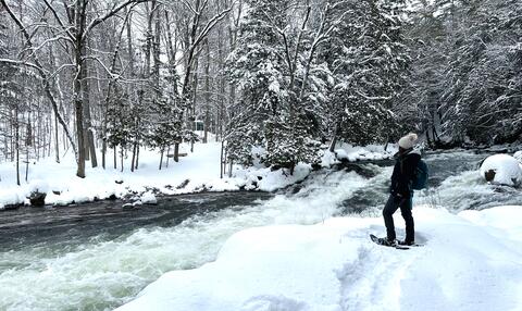 A woman stands in snowshoes overlooking river rapids.