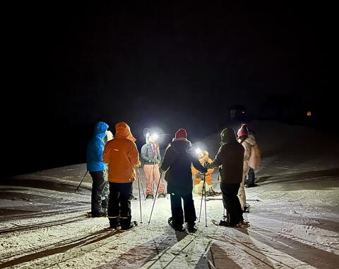 Snowshoers gather at night using flashlights in the snow.